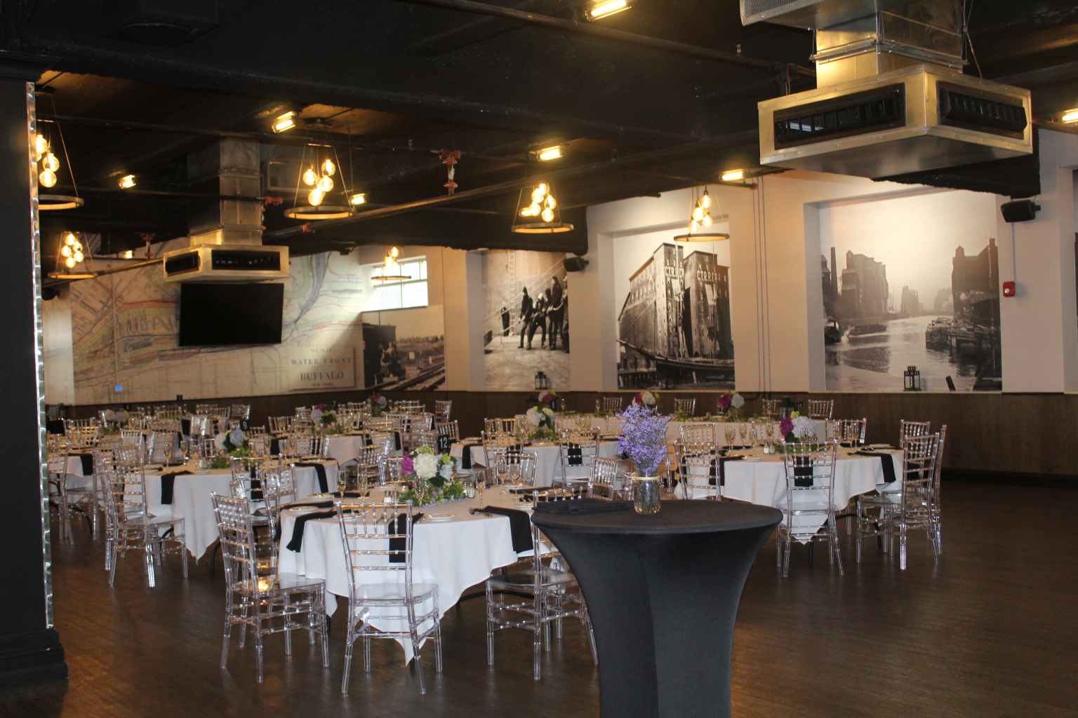 A private banquet room set with round tables, white linens, floral centerpieces and clear chiavari chairs