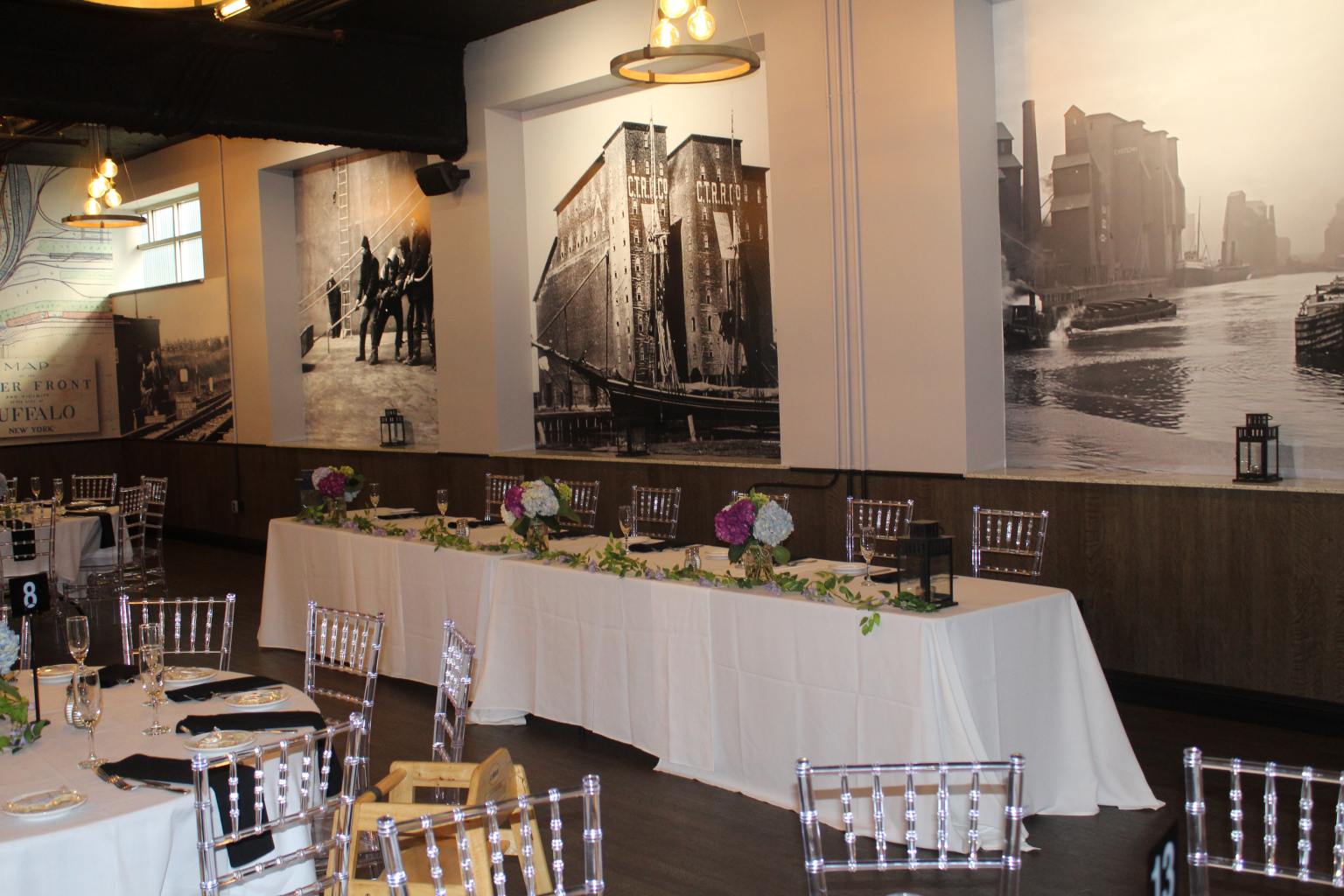 Head table set with white linen, greenery and clear chiavari chairs in front of the Ganson Street murals